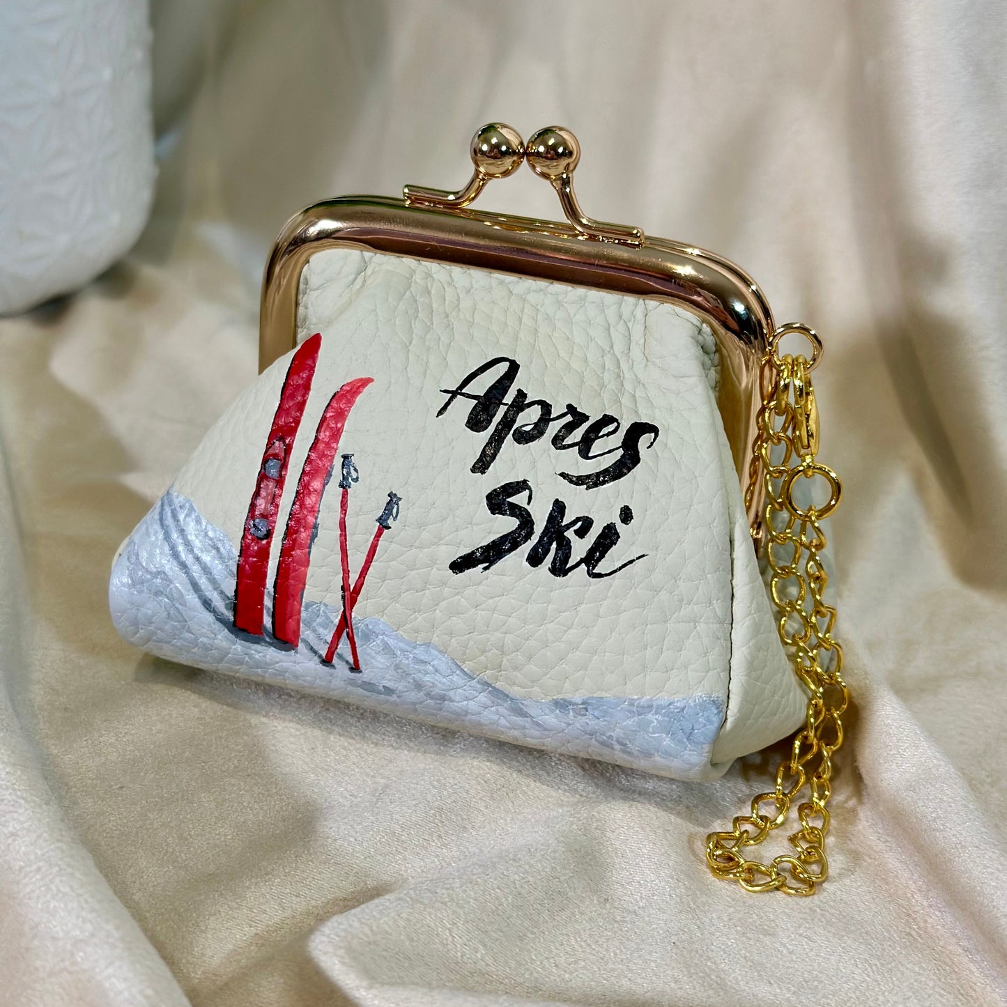 Small white purse with 'Apres Ski' text and skis design on a beige fabric background