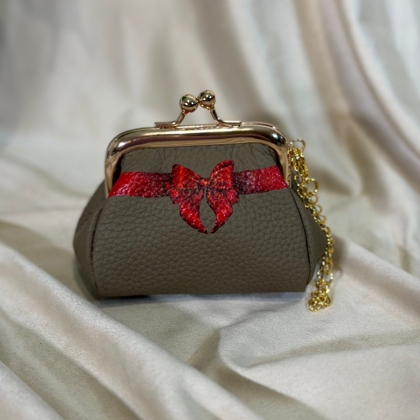 Small gray purse with a red bow and gold chain on a white fabric background