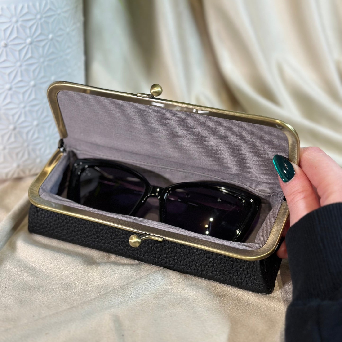 Black sunglasses in a textured black case held by a hand with green nail polish.