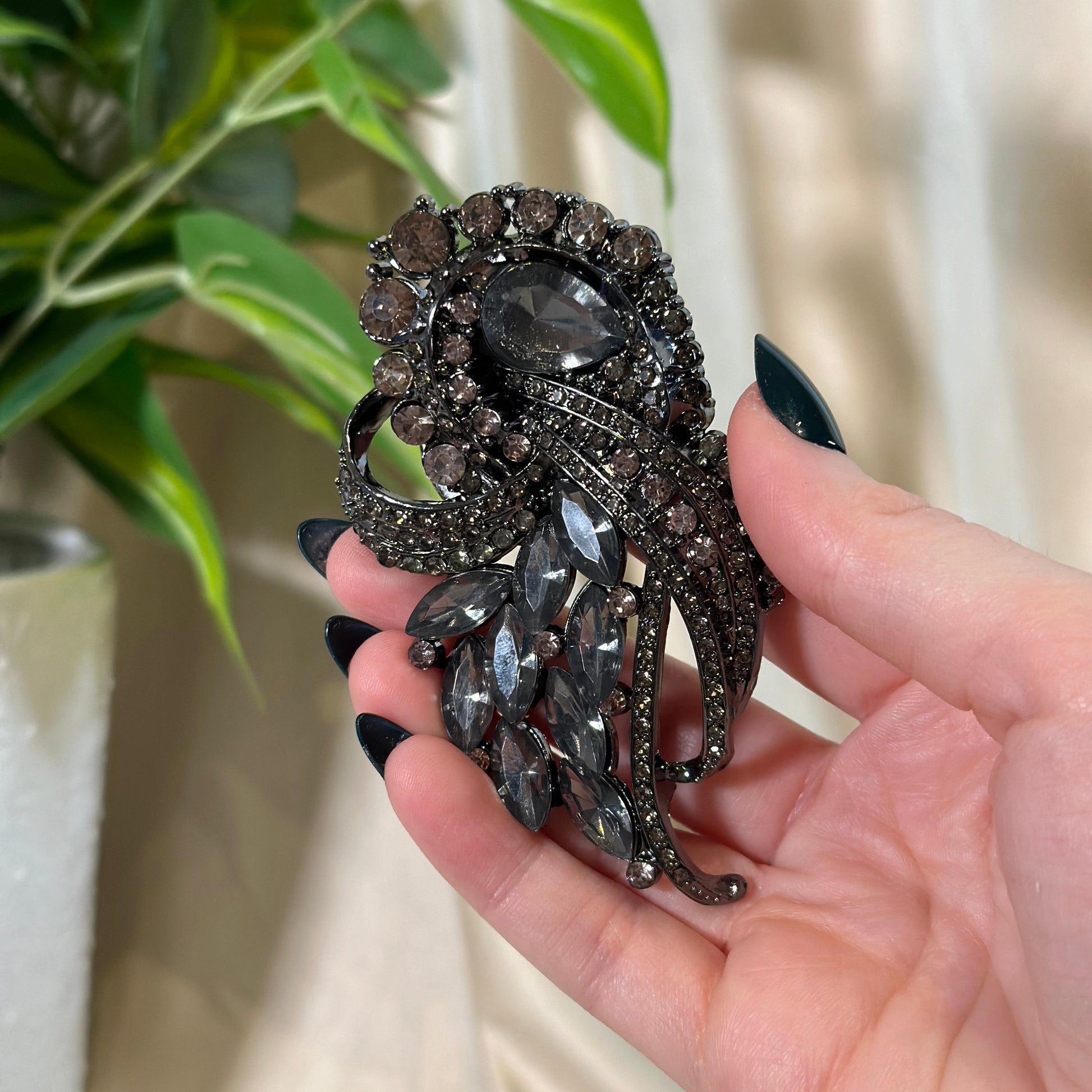 Hand holding a decorative brooch with intricate design and gemstones, blurred plant and chair in the background