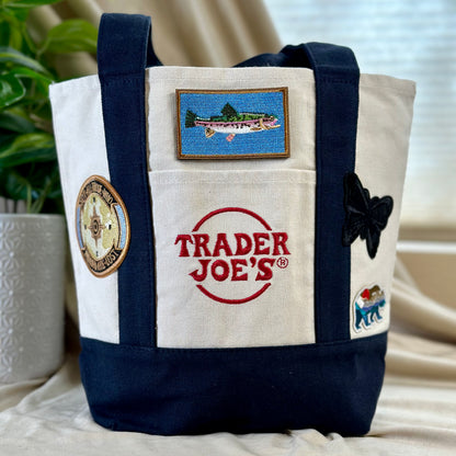 A canvas tote bag with navy blue trim and handles, featuring embroidered patches including a fish patch, a compass patch, a black butterfly patch, and a Trader Joe's logo patch on the front pocket.