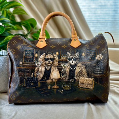 A dark brown monogram handbag with tan leather handles, featuring a detailed illustration of a cat and dog dressed in luxury fashion at a table, set against a background of beige curtains and a green plant.