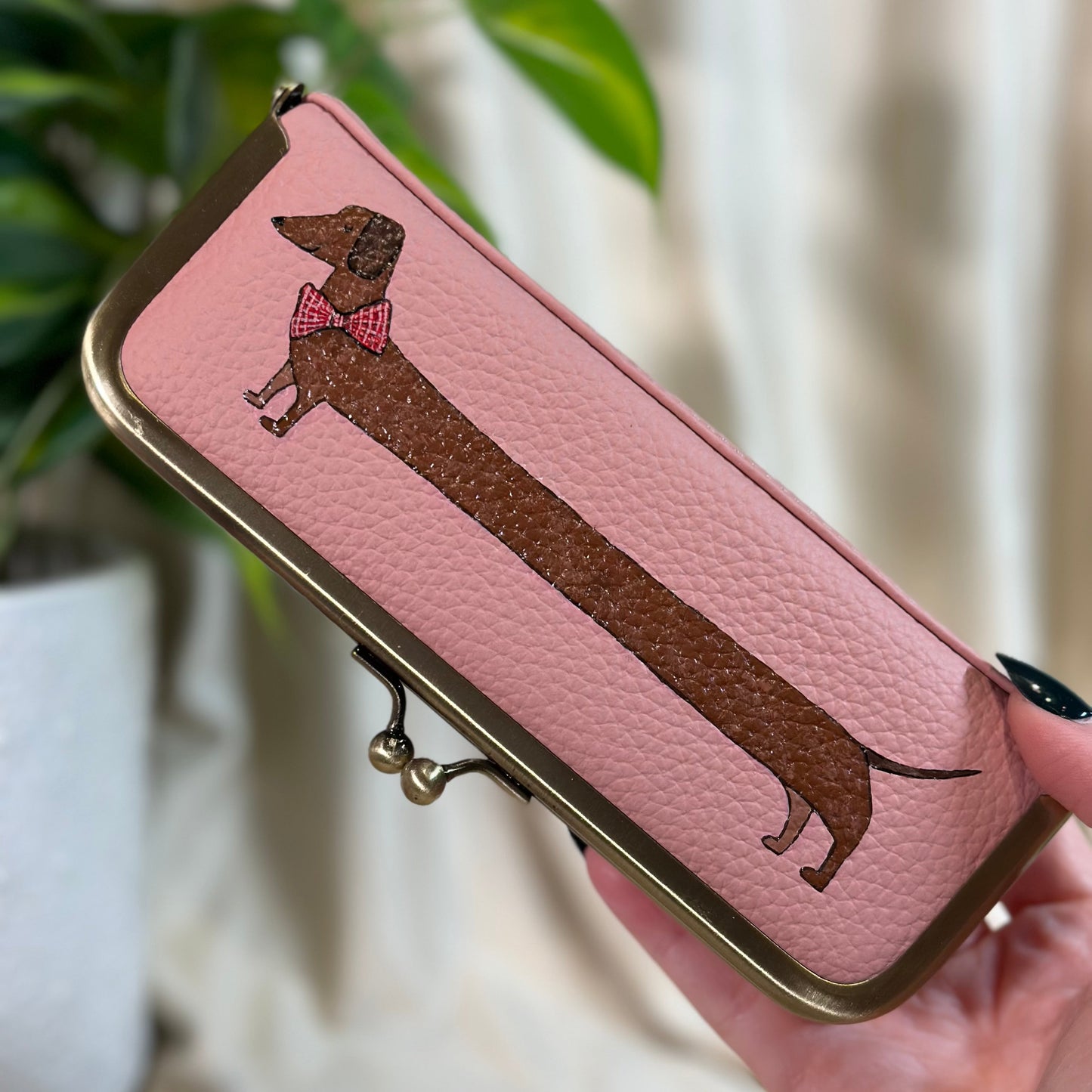 Pink wallet with dachshund design held by a hand, blurred plant and curtain background
