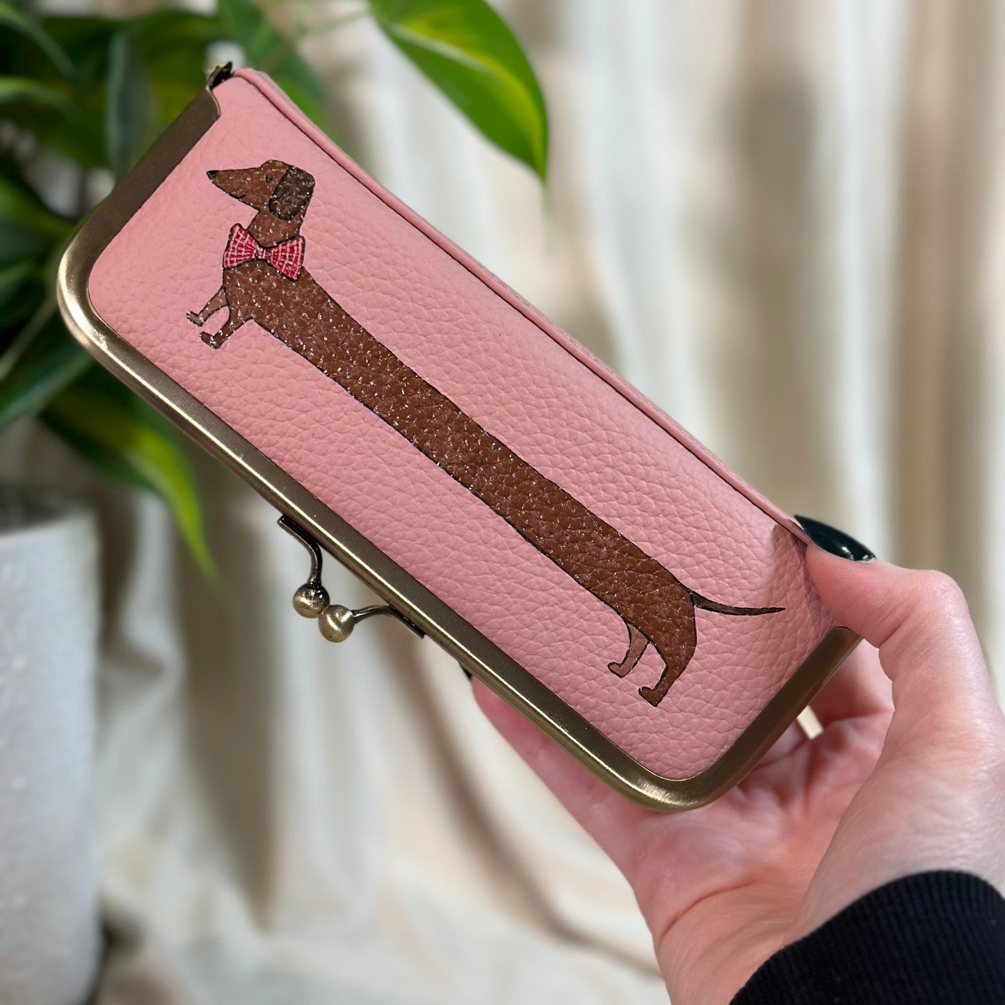 Pink pouch with dachshund design held by a hand, plant in the background