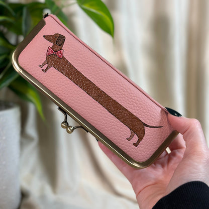 Pink pouch with dachshund design held by a hand, plant in the background