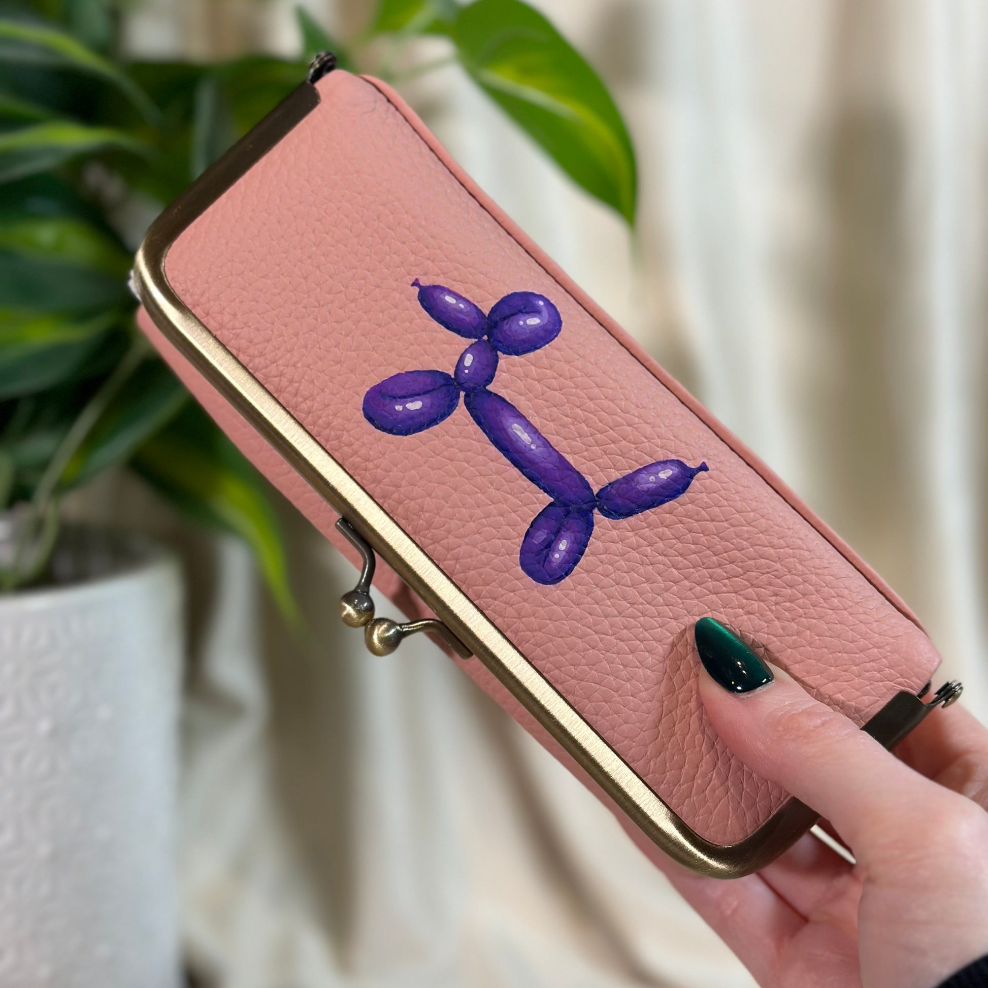 Pink phone case with purple balloon dog design held by a hand, plant in background