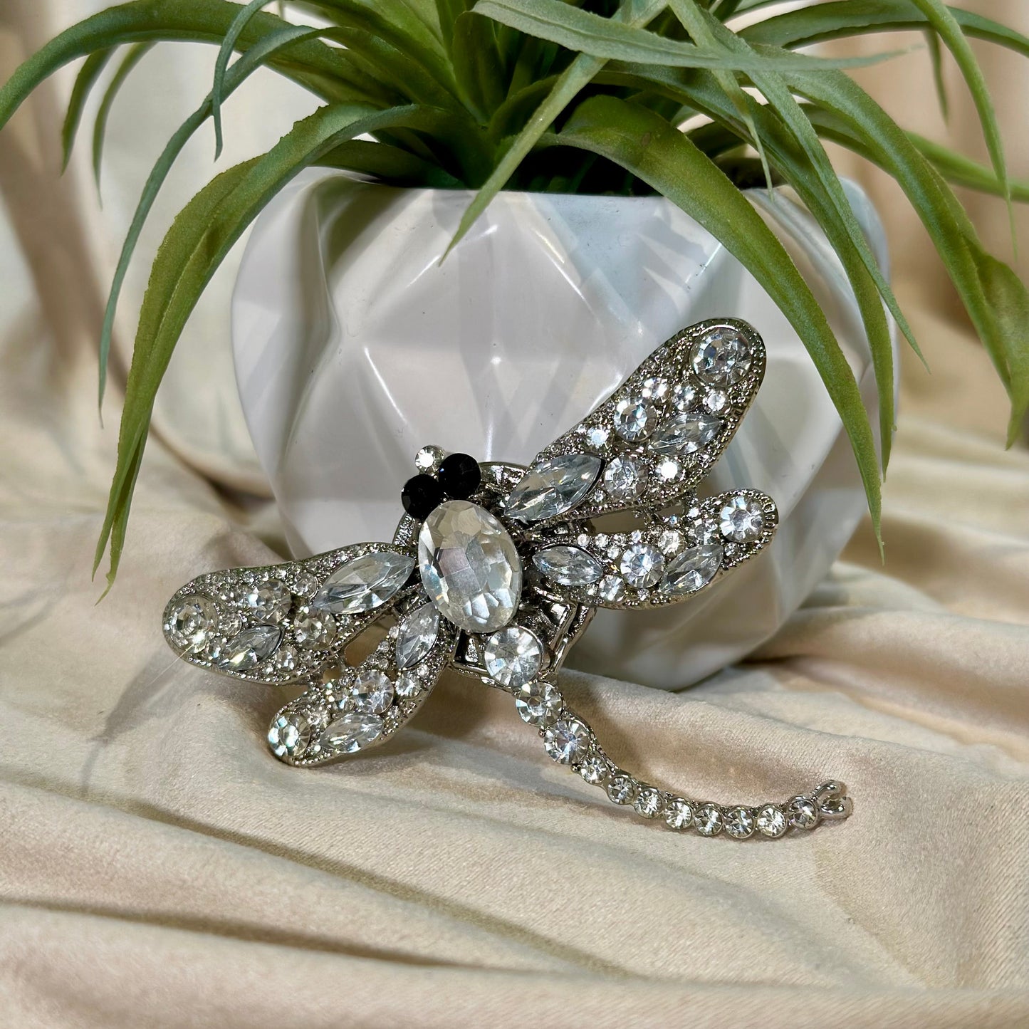 Silver dragonfly brooch with clear gemstones on a beige surface with a plant in the background