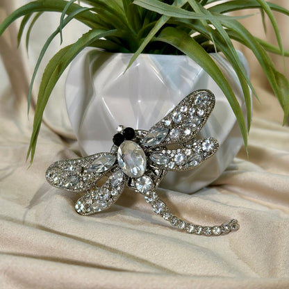 Silver dragonfly brooch with clear gemstones on a beige surface with a plant in the background