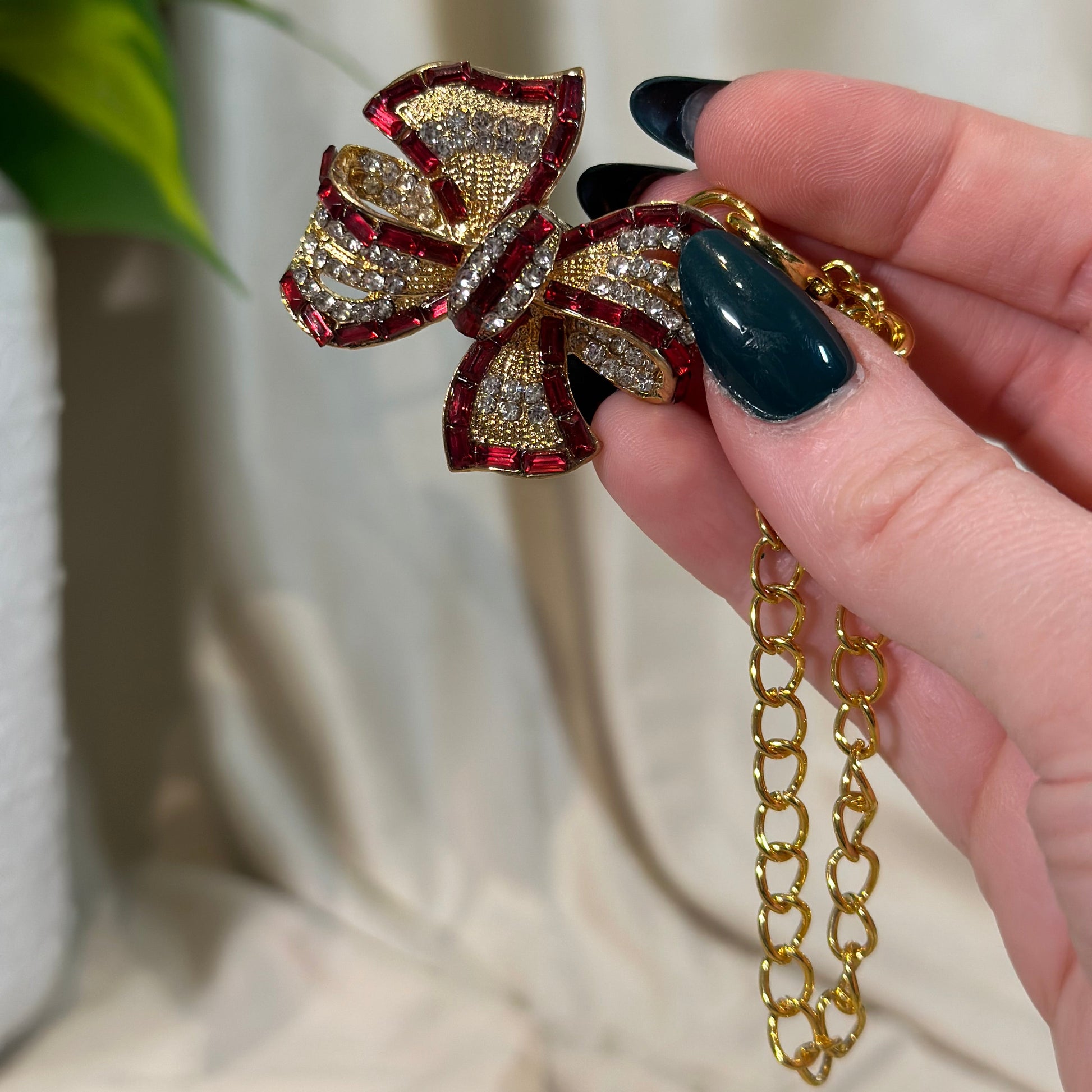 Gold and red bow-shaped pendant on a gold chain held by a hand with dark nail polish.