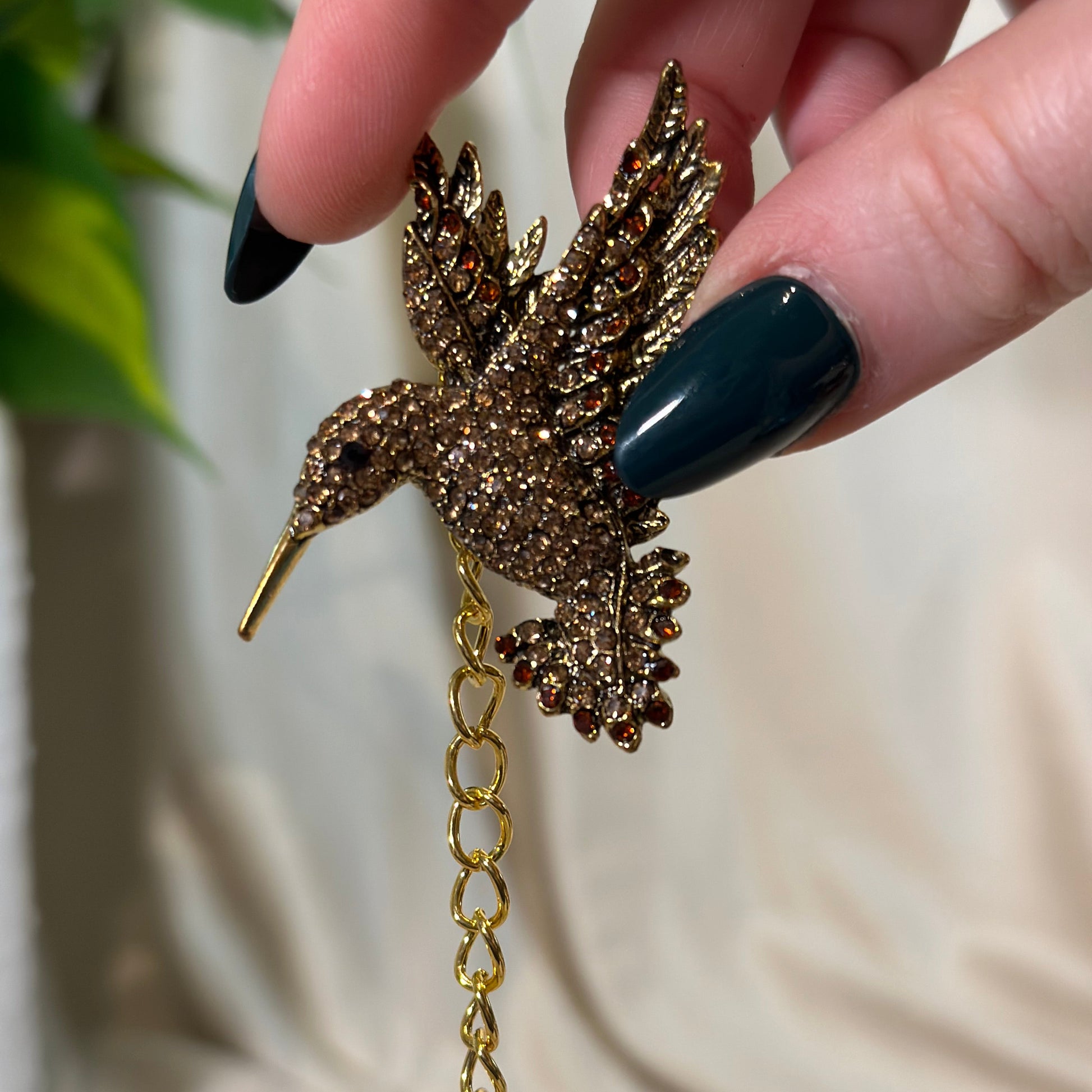 Gold bird-shaped brooch held by a hand with dark nail polish against a neutral background
