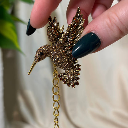 Gold bird-shaped brooch held by a hand with dark nail polish against a neutral background