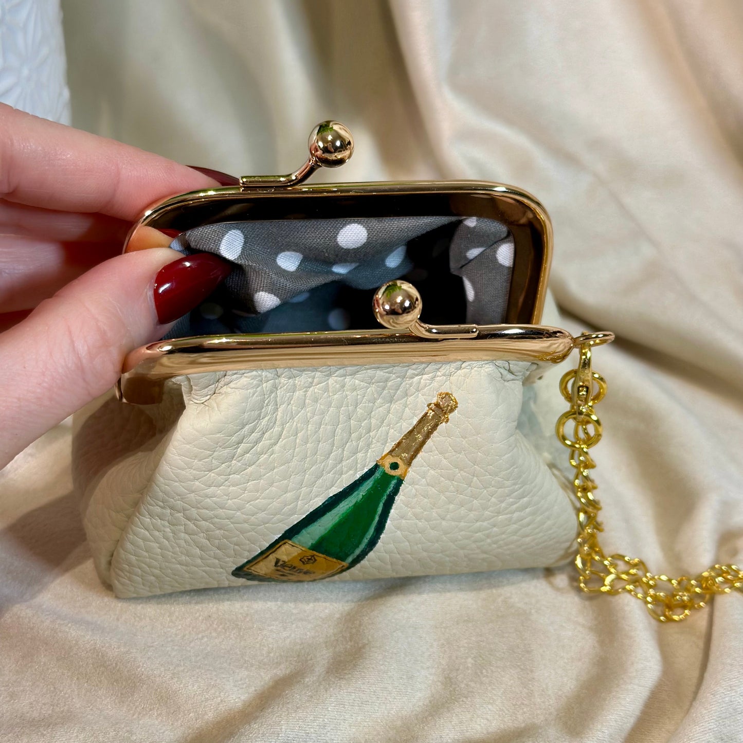 Small handbag with champagne bottle design held by a hand on a beige fabric background