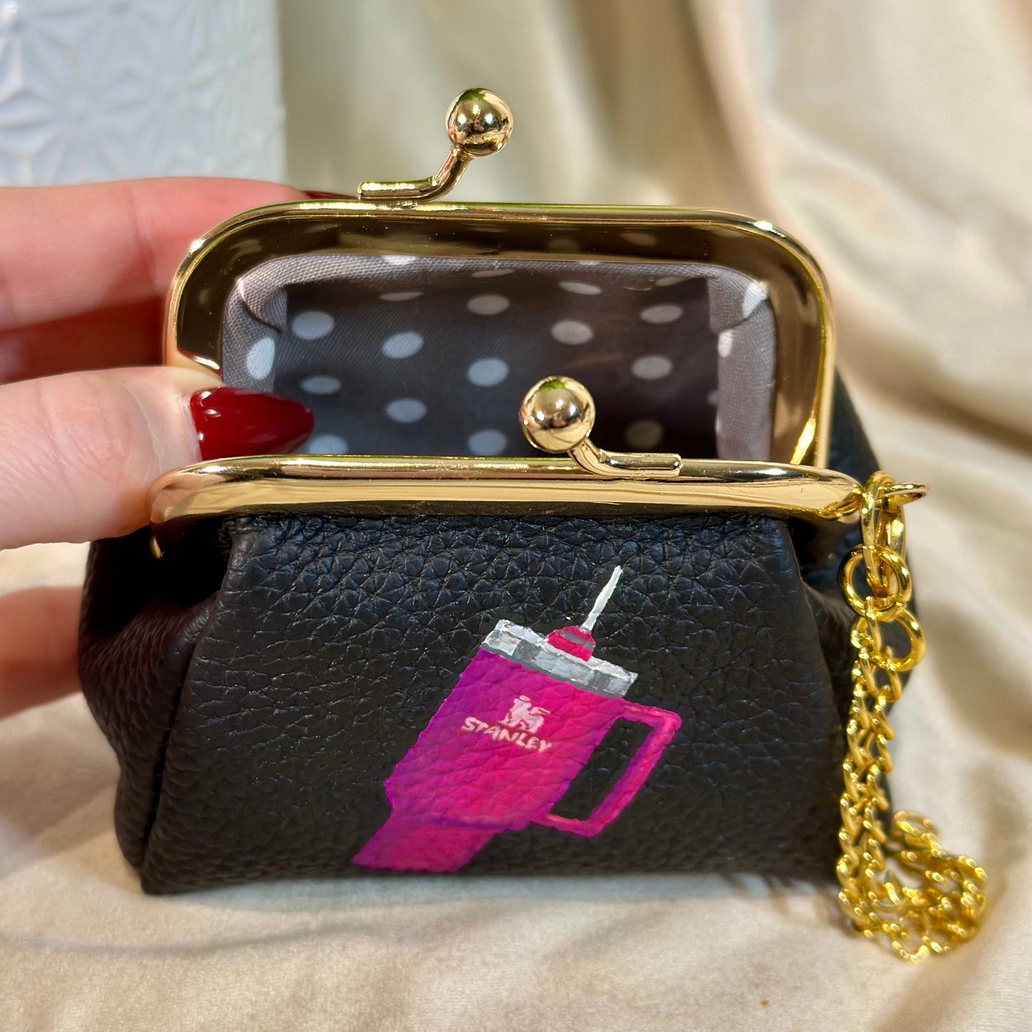 Black leather coin purse with gold clasp and chain, featuring a pink drink design, held by a hand.