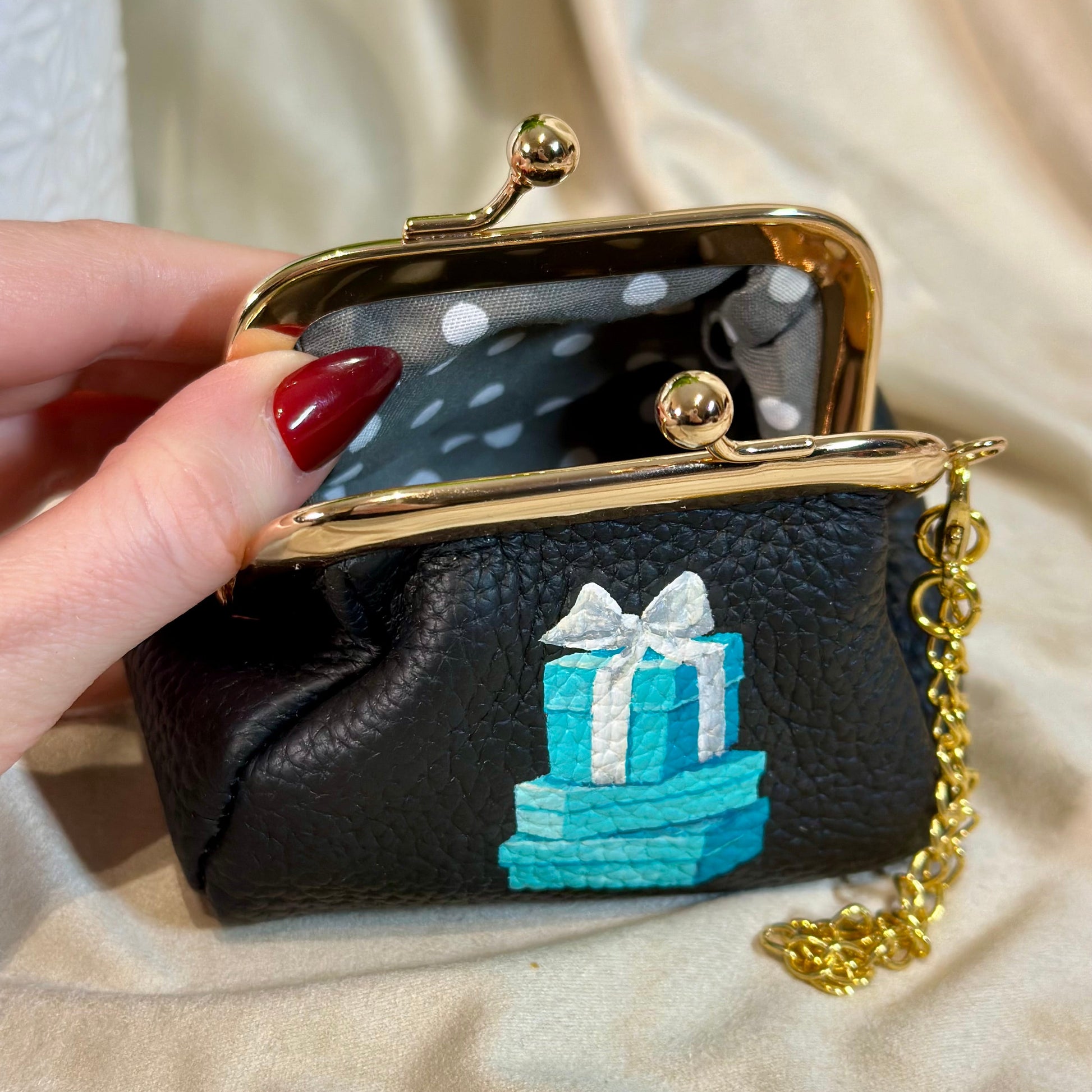 Black purse with blue gift box design held by a hand on a beige fabric background