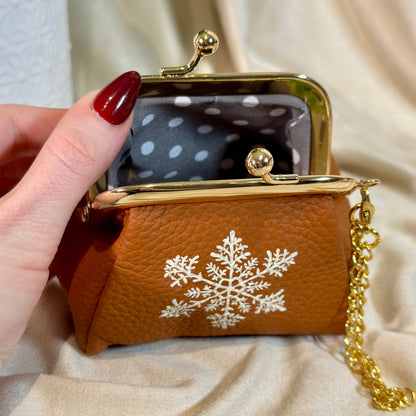 Brown clutch with gold chain held by a hand, featuring a white snowflake design on a beige fabric background.
