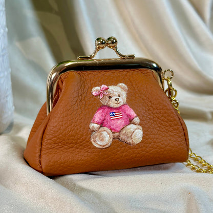 Brown leather purse with a teddy bear embroidered on it, placed on a white fabric background.