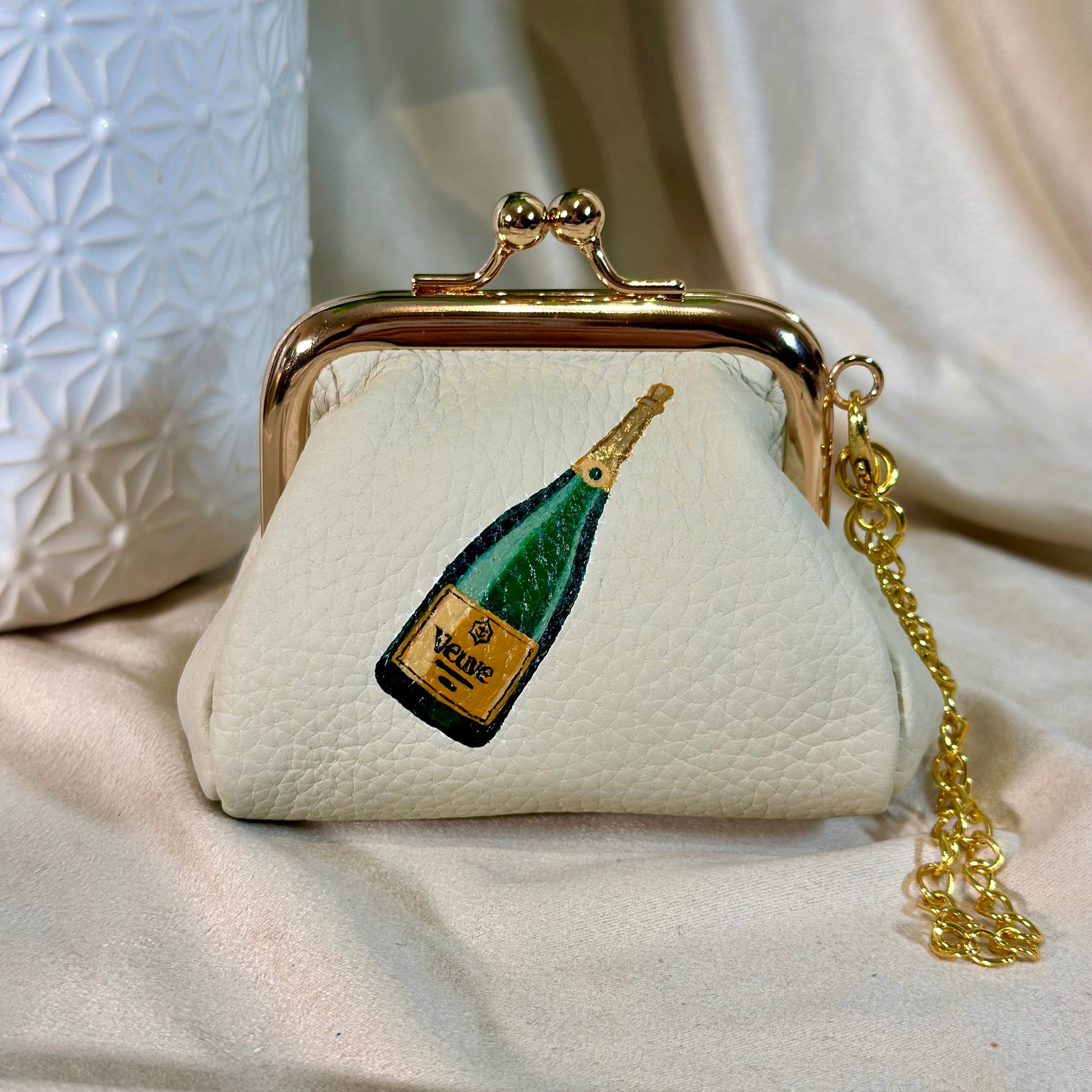 Small cream-colored purse with champagne bottle design and gold chain on a light fabric background