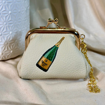 Small cream-colored purse with champagne bottle design and gold chain on a light fabric background