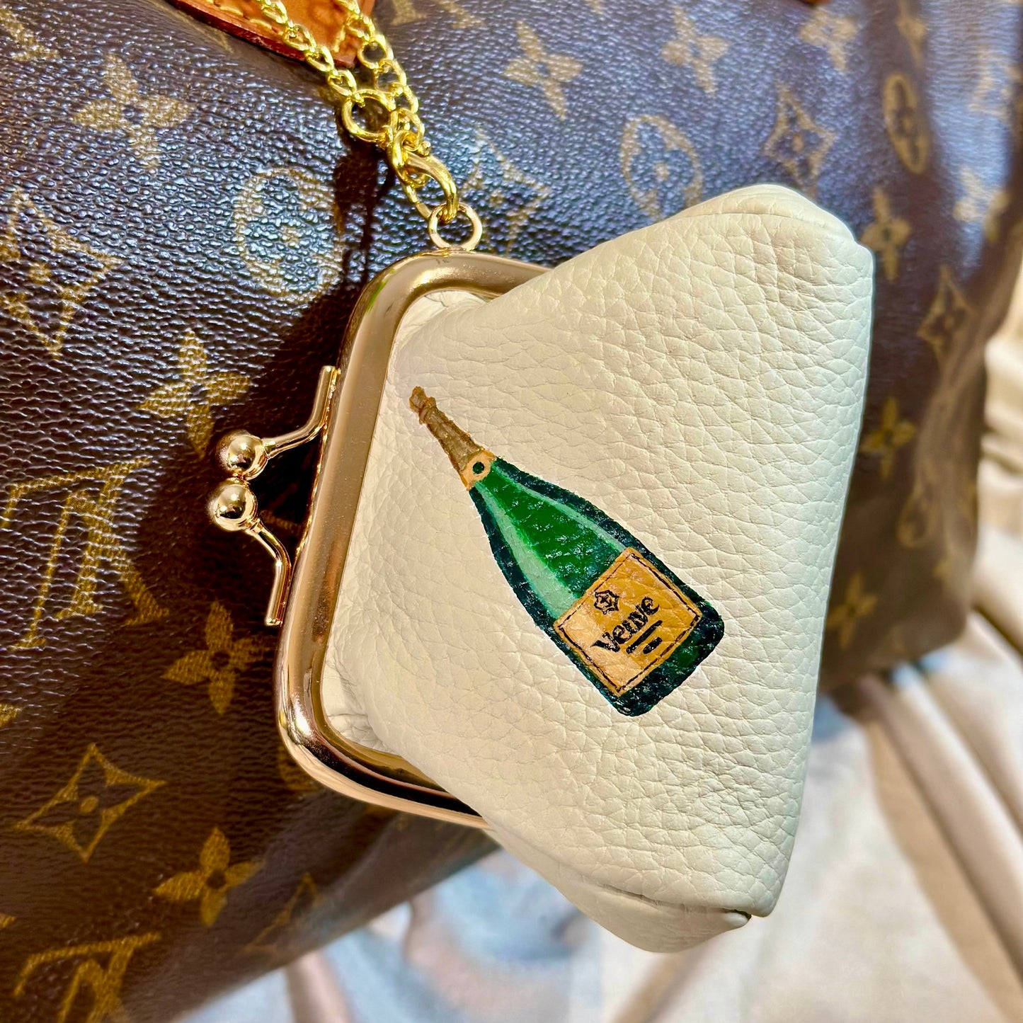 Small cream-colored purse with a green bottle design in front of a brown Louis Vuitton bag.