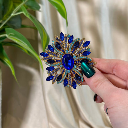 Hand holding a decorative brooch with blue and gold details against a neutral background