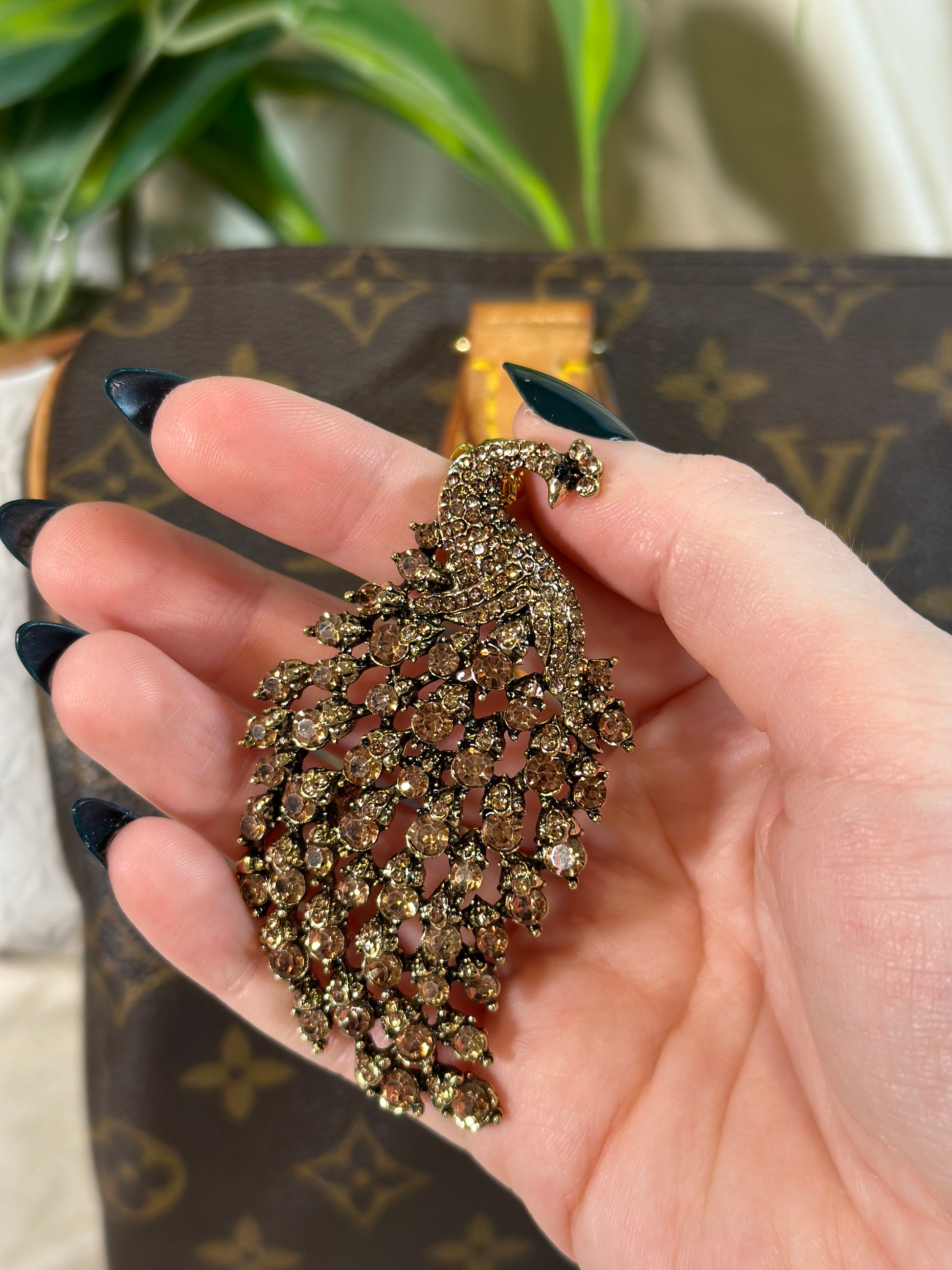 Hand holding a decorative brooch with a blurred Louis Vuitton bag in the background