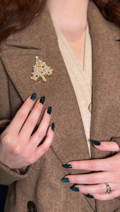 Person wearing a brown coat with a decorative brooch, holding the coat with green nail polish.