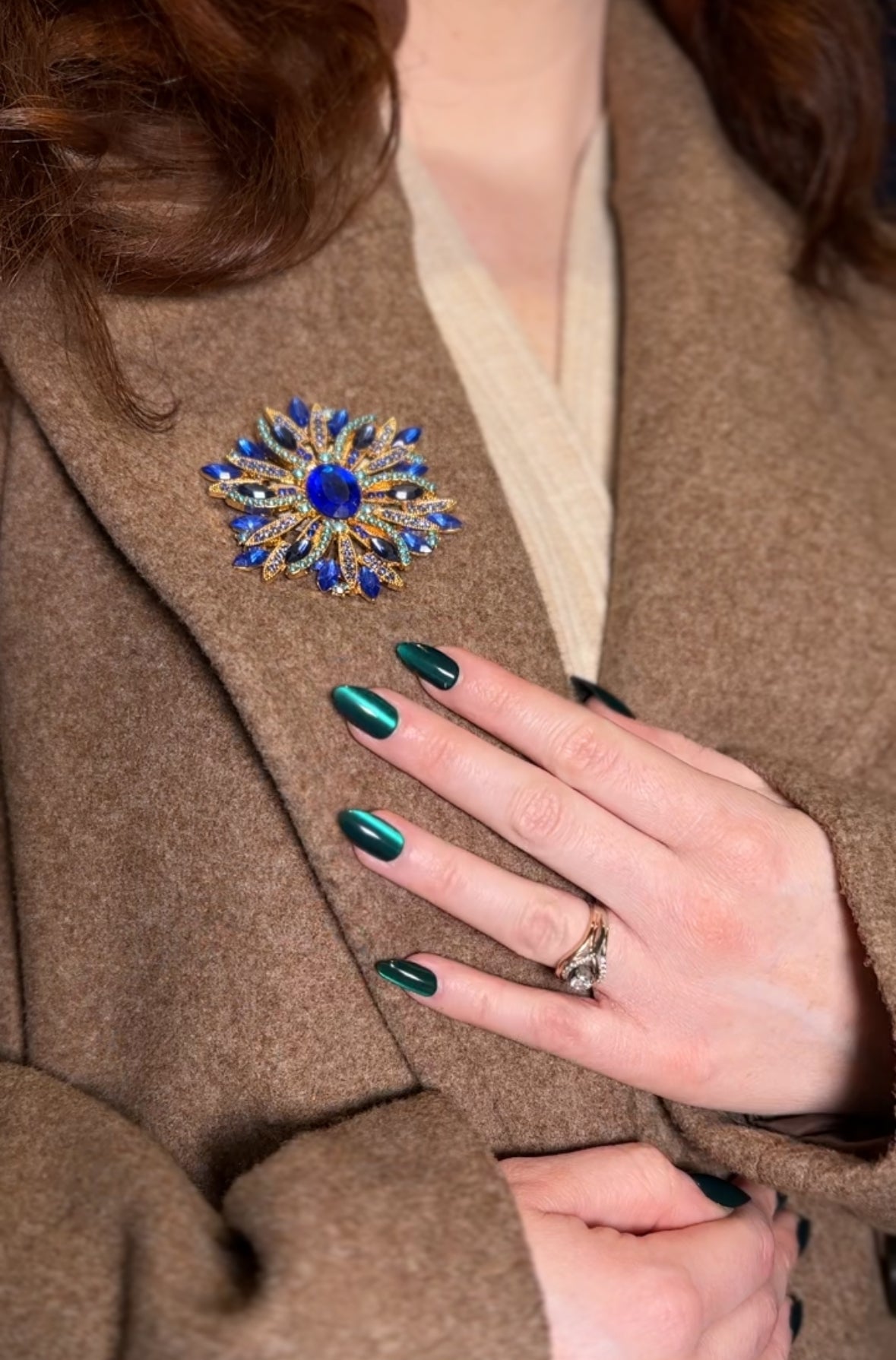 Close-up of a person wearing a decorative brooch on a brown coat with green nail polish.