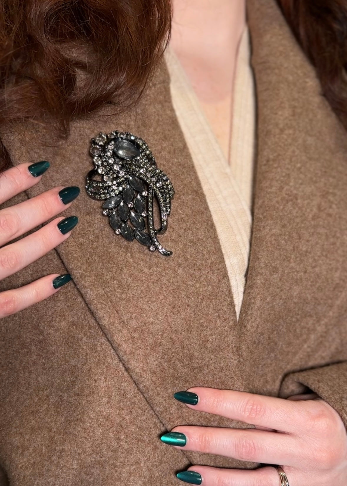 Person wearing a decorative brooch on a brown coat with green nail polish.