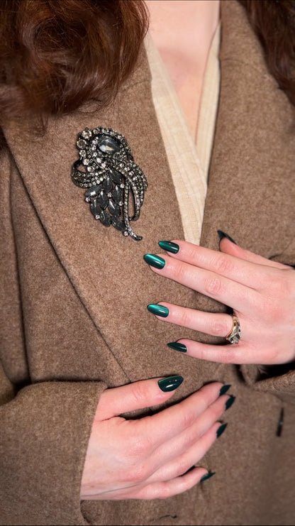 Person wearing a brown coat with a decorative brooch, showing hands with green nail polish.