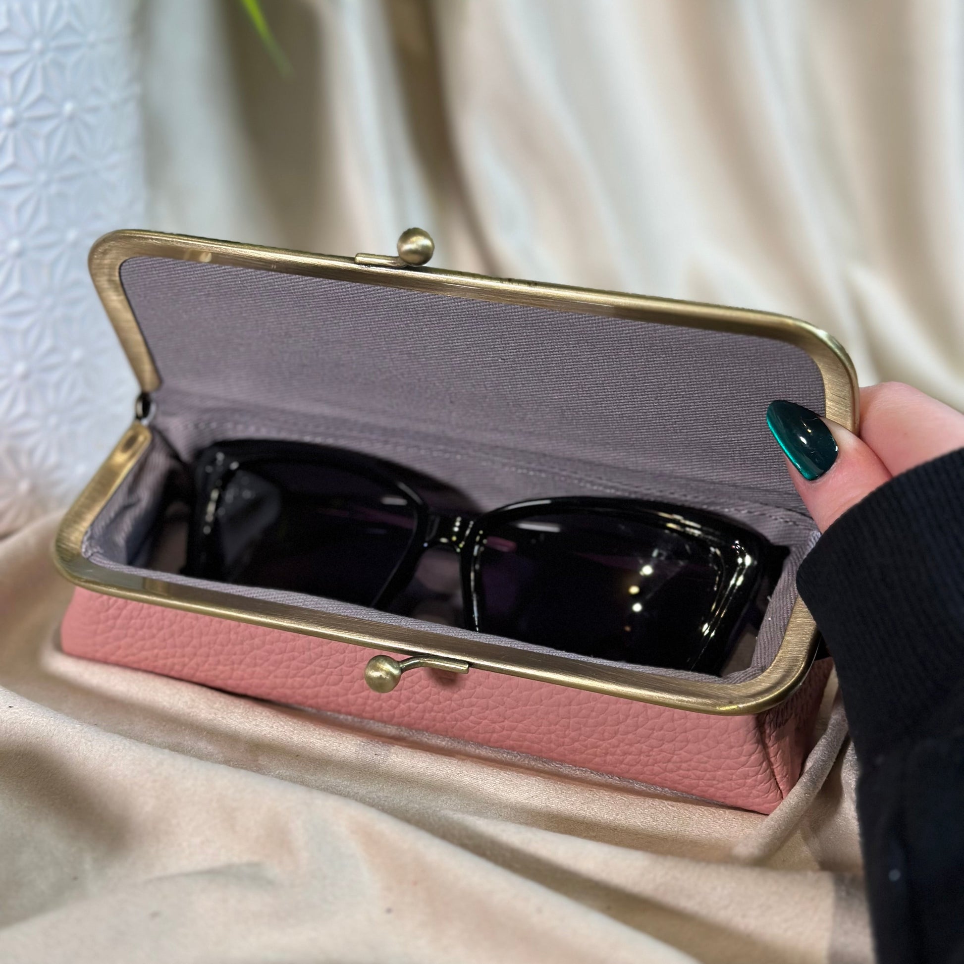 Pink sunglasses case with sunglasses inside, held by a hand against a neutral background
