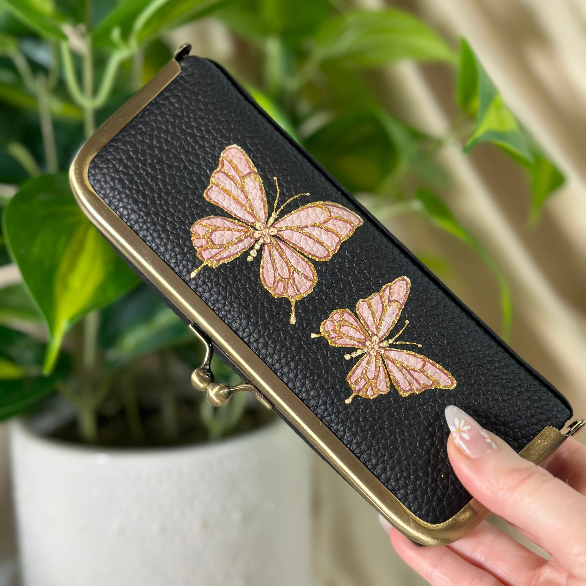 Black pouch with butterfly designs held by a hand in front of a plant