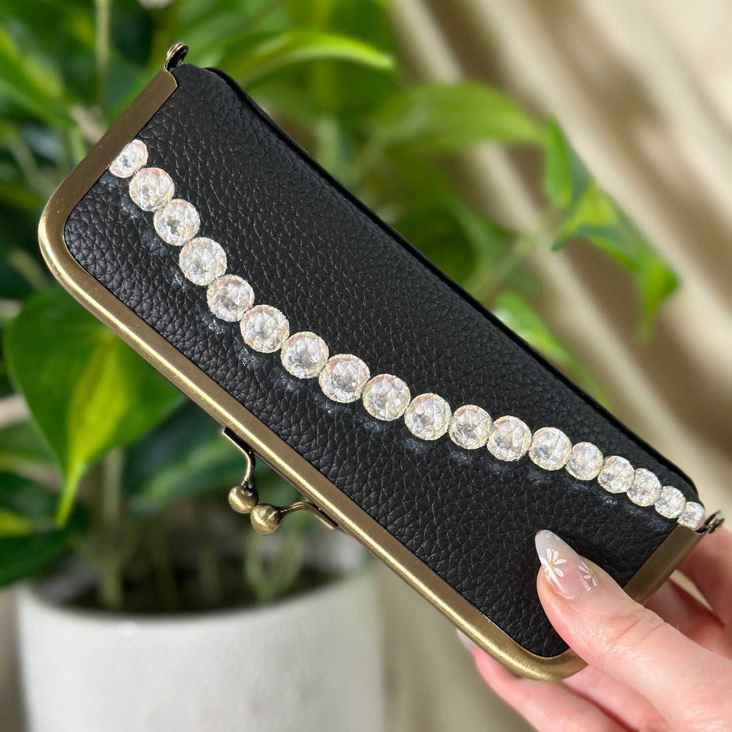 Black clutch with pearl embellishments held by a hand in front of a plant