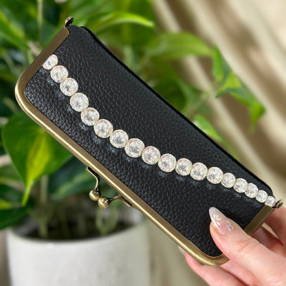 Black clutch with pearl embellishments held by a hand in front of a plant