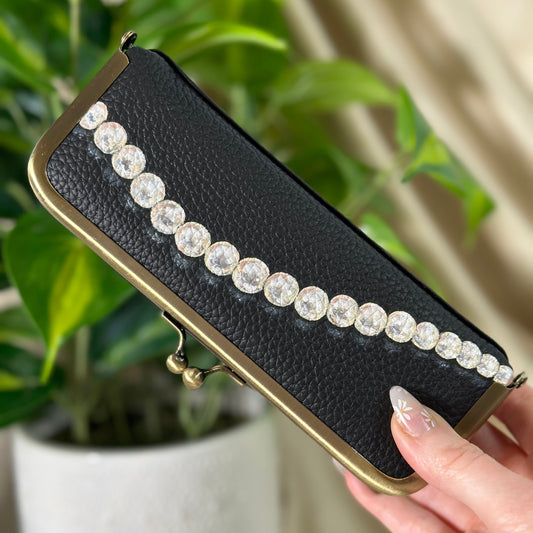 Black clutch with pearl embellishments held by a hand in front of a plant