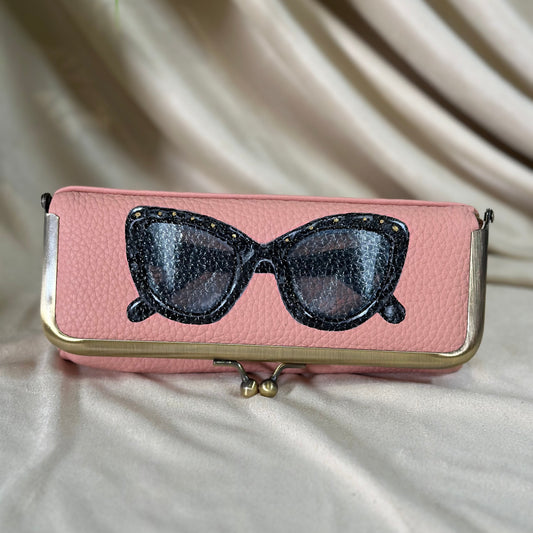 Pink clutch with black cat-eye sunglasses design on a beige fabric background