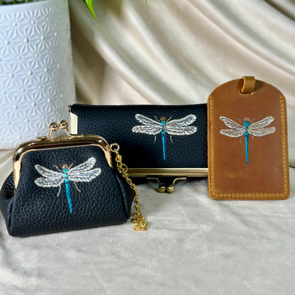 Black clutch with dragonfly design, stand, and brown luggage tag on a light background
