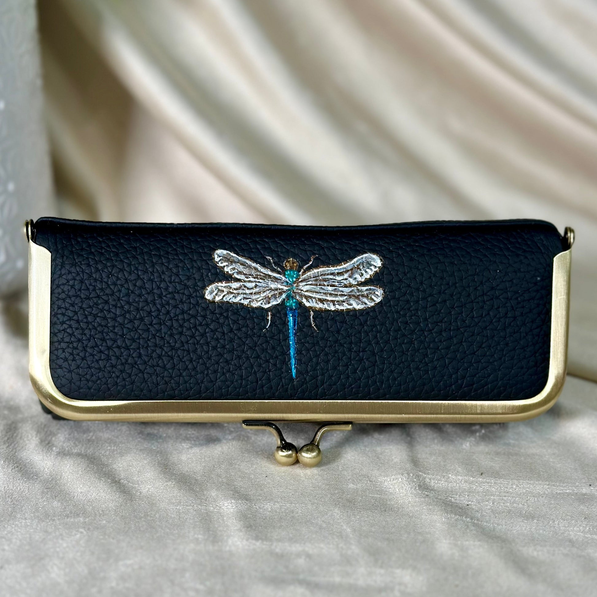 Black pouch with gold frame and embroidered dragonfly design on a light gray background