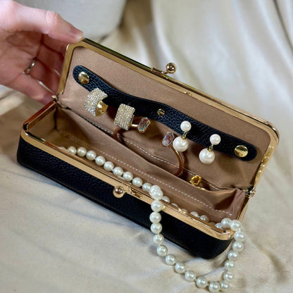 A black faux-leather jewelry organizer case with a gold-tone frame, held open by a hand. Inside, a black leatherette organizer strip holds rings, and a string of white faux pearls is draped over the front.