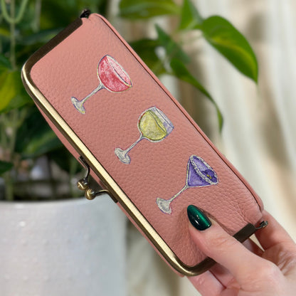 Pink wallet with colorful wine glass designs held by a hand in front of a plant.