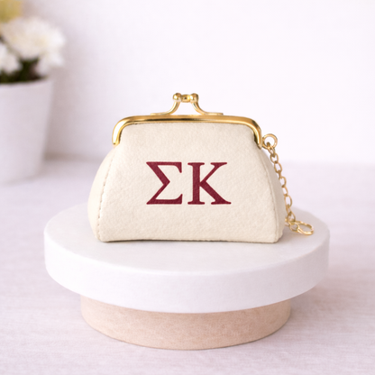 Small cream-colored purse with gold frame and red 'ΣK' letters on a white surface.