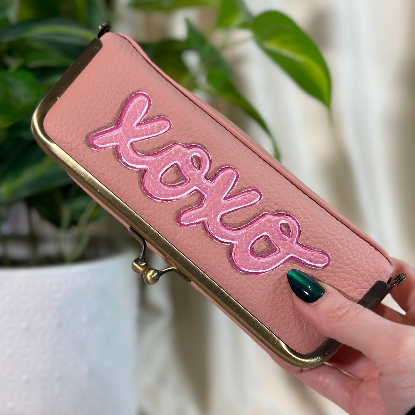 Pink clutch with 'xoxo' embroidery held by a hand in front of a plant.