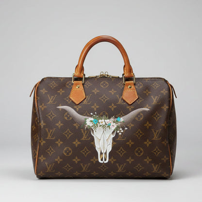 Designer handbag with a cow skull and floral design on a wooden bench with plants in the background