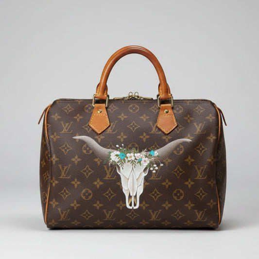 Designer handbag with a cow skull and floral design on a wooden bench with plants in the background